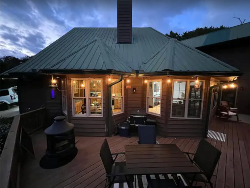 Cozy cabin with a green metal roof, surrounded by a deck and outdoor seating, illuminated by string lights at dusk.