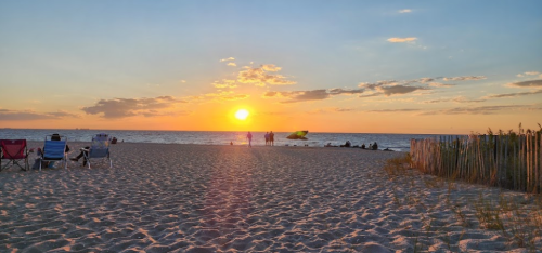 A serene beach scene at sunset, with people walking along the shore and chairs set up on the sand.