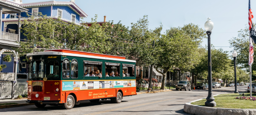 A colorful trolley bus drives down a tree-lined street with historic buildings and parked cars in the background.