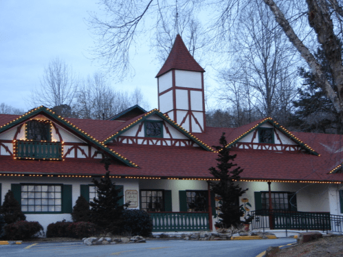 A charming building with a steep roof, decorative lights, and a tower, surrounded by trees in a twilight setting.