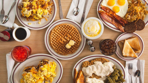 A spread of breakfast dishes including eggs, bacon, waffles, hash browns, and toast on a wooden table.
