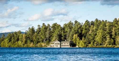 A serene lake scene featuring a large house surrounded by lush green trees under a blue sky with fluffy clouds.