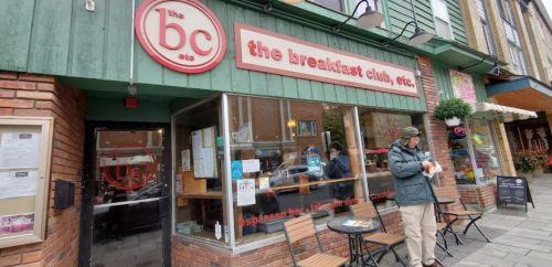 A cozy café called "the breakfast club, etc." with outdoor seating and a customer standing outside.