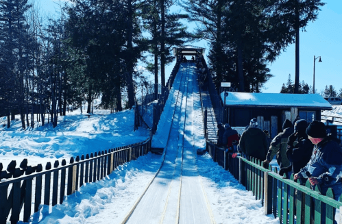 A snowy hill with a wooden slide, surrounded by trees, and people waiting in line on a sunny winter day.