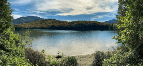 A serene lake surrounded by colorful autumn foliage and mountains under a blue sky with wispy clouds.