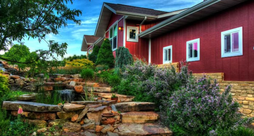 A red house surrounded by lush greenery, flowers, and a small pond with a waterfall, under a blue sky.