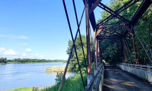 A scenic view of a river alongside an old metal bridge, surrounded by lush greenery and a clear blue sky.
