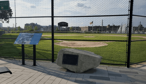 A baseball field behind a fence, with a stone monument and informational plaque in the foreground.
