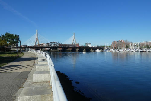 A scenic view of a river with a bridge, boats, and a clear blue sky, alongside a walking path and greenery.