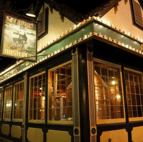 A cozy Irish pub with a green exterior, decorative lights, and large windows showcasing brewing equipment inside.