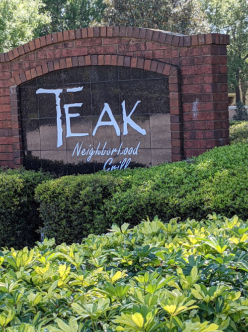 Sign for "TEAK Neighborhood Grill" surrounded by green bushes and plants.