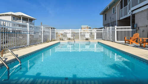 A clear blue swimming pool surrounded by a white fence and lounge chairs under a bright blue sky.