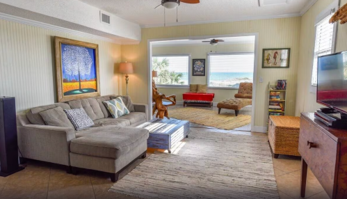 Cozy living room with a gray sofa, colorful artwork, and a view of the beach through large windows.