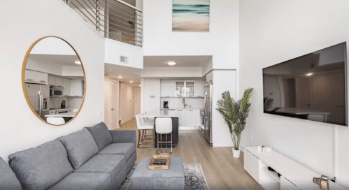 Modern living room with a gray sofa, large mirror, TV, and open kitchen in a bright, airy space.