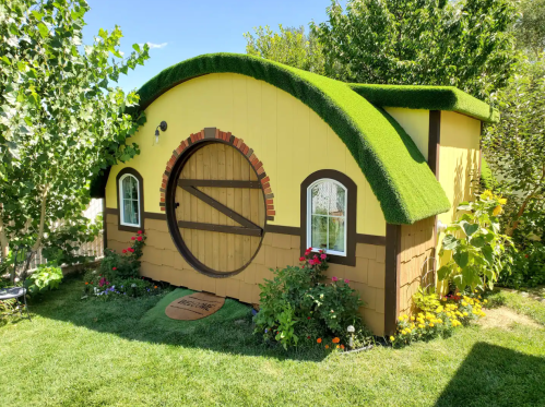 A whimsical yellow hobbit-style shed with a rounded green roof, surrounded by colorful flowers and greenery.