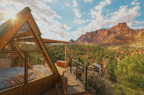 A modern A-frame cabin overlooks a lush valley and mountains under a bright sky with scattered clouds.