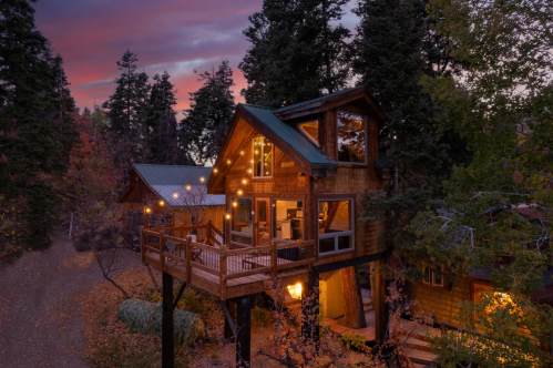 A cozy wooden cabin surrounded by trees, featuring a deck with lights, set against a colorful sunset sky.