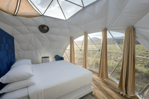 A cozy, modern geodesic dome interior with a bed, large windows, and scenic views of nature outside.