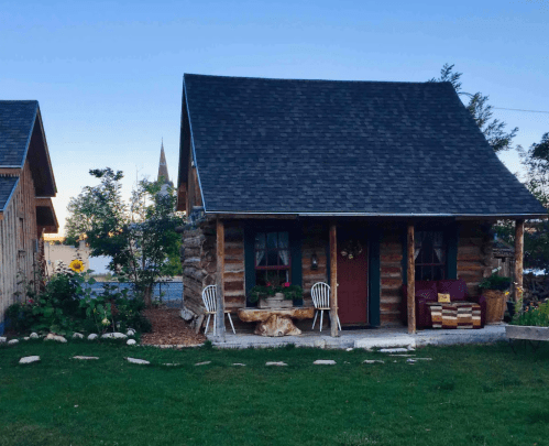 A cozy log cabin with a porch, surrounded by greenery and flowers, under a clear blue sky.
