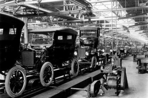 Historic black-and-white photo of early automobiles on an assembly line in a factory.