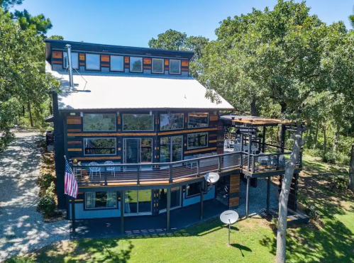 A modern two-story house with large windows, a balcony, and a spacious yard surrounded by trees.
