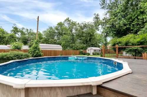 A round above-ground pool surrounded by a wooden deck and lush greenery in a backyard setting.
