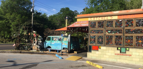 A colorful pizza shop with stained glass windows and a vintage blue van parked outside, surrounded by greenery.