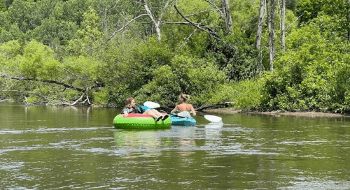 Two people float on colorful inner tubes in a calm river surrounded by lush greenery.