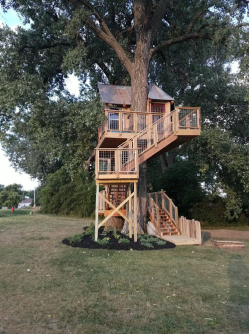 A wooden treehouse with a metal roof, featuring a staircase and balcony, nestled in a large tree in a grassy area.