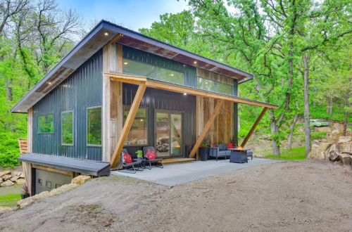 Modern cabin with large windows, wooden accents, and a patio, surrounded by lush greenery and rocky terrain.