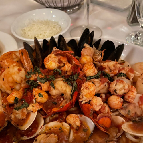 A seafood dish featuring shrimp, lobster, clams, and mussels, garnished with herbs, served with a side of grated cheese.