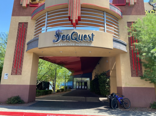 Entrance of SeaQuest Interactive Aquarium, featuring a large sign and a bike parked nearby, surrounded by greenery.