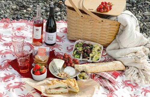 A picnic spread on a blanket featuring wine, salads, sandwiches, fruits, and a woven basket.