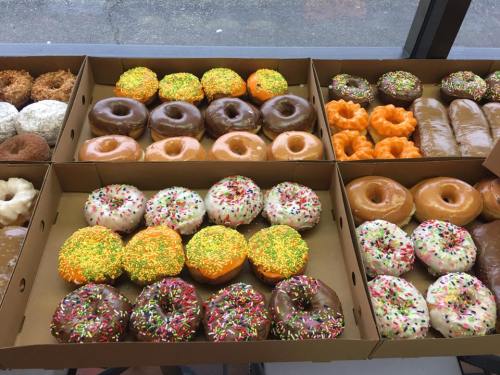 A variety of colorful donuts in boxes, featuring sprinkles, chocolate, and orange icing.