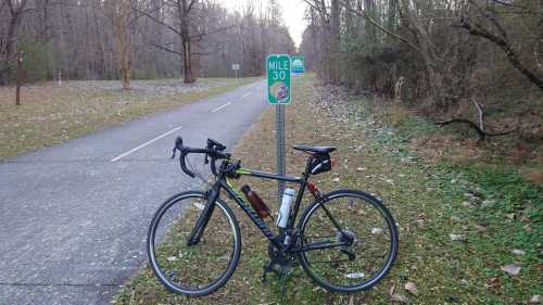 A black bicycle parked next to a mile marker sign on a tree-lined path, indicating mile 30.