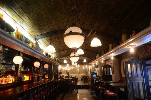 A cozy bar with wooden beams, warm lighting, and a long counter lined with stools and bottles.