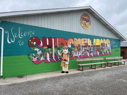 A costumed character stands in front of a colorful mural that says "Welcome to Shipshewana." Benches are nearby.