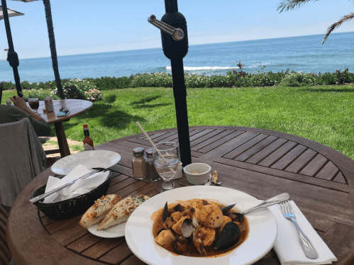 A plate of seafood pasta on a wooden table with a view of the ocean and green grass in the background.