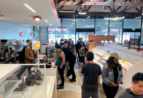 A busy café interior with a line of customers at the counter and people browsing the display cases.