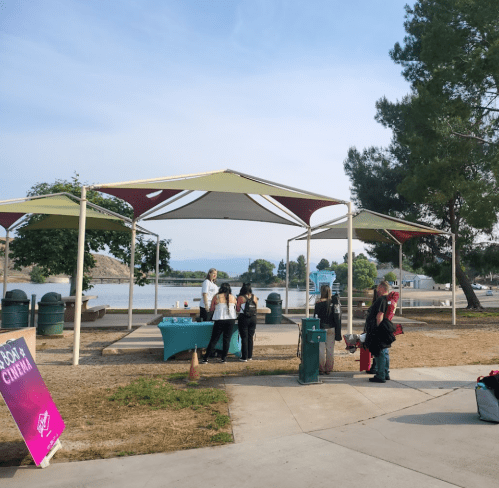 A group of people gathers under a shaded pavilion by a lake, with trees and a clear sky in the background.