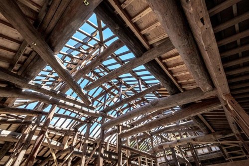 View of a wooden roof structure with exposed beams and a clear blue sky visible through the openings.
