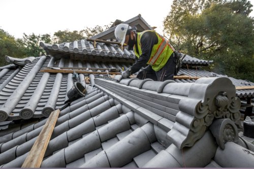 A worker in a hard hat and safety vest repairs a traditional tiled roof, surrounded by trees and architectural details.