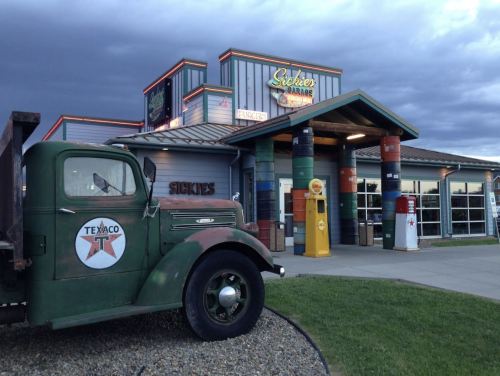 A vintage green truck parked outside a colorful gas station with retro signage and barrels in front.