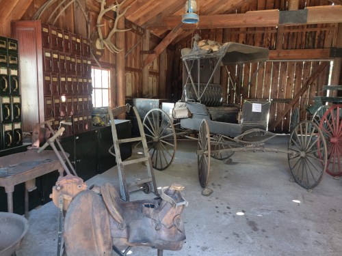 Interior of a rustic barn displaying vintage carriages and tools, with wooden beams and shelves in the background.