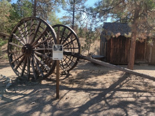 A large wooden wheel structure beside a small wooden shed in a forested area, with a sign nearby.