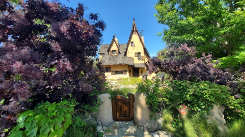 A whimsical, fairy-tale style house surrounded by lush greenery and vibrant purple foliage under a clear blue sky.