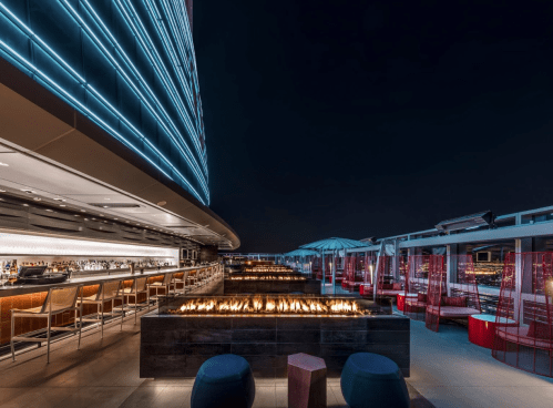 Rooftop bar at night with glowing blue lights, fire pits, and modern seating overlooking a city skyline.