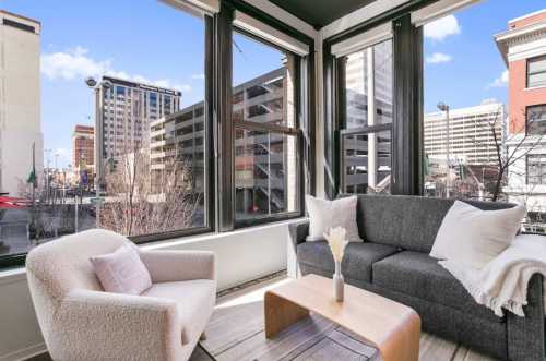 Bright, modern living space with large windows, a gray sofa, a cozy chair, and a wooden coffee table overlooking a cityscape.