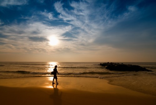 A silhouette of a person walking along a beach at sunset, with waves gently lapping at the shore.