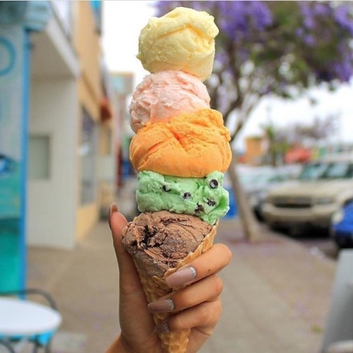 A hand holding a tall ice cream cone with five colorful scoops: chocolate, mint, orange, strawberry, and vanilla.